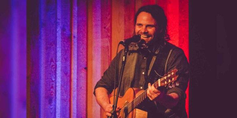 Musiker Keller Steff sitzt mit Gitarre und Mikrofon auf einer Bühne und lächelt